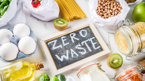 Foods including eggs, pasta, oil, fruit, pulses arranged on a table. In the centre,  a chalkboard bears the slogan "Zero waste".