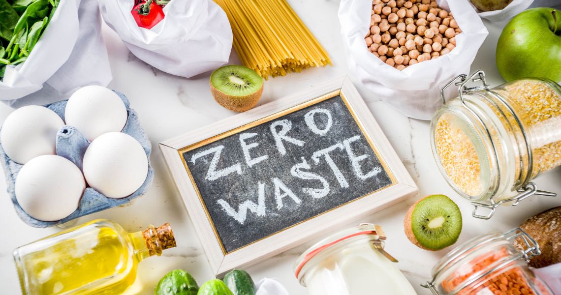 Foods including eggs, pasta, oil, fruit, pulses arranged on a table. In the centre, a chalkboard bears the slogan "Zero waste".