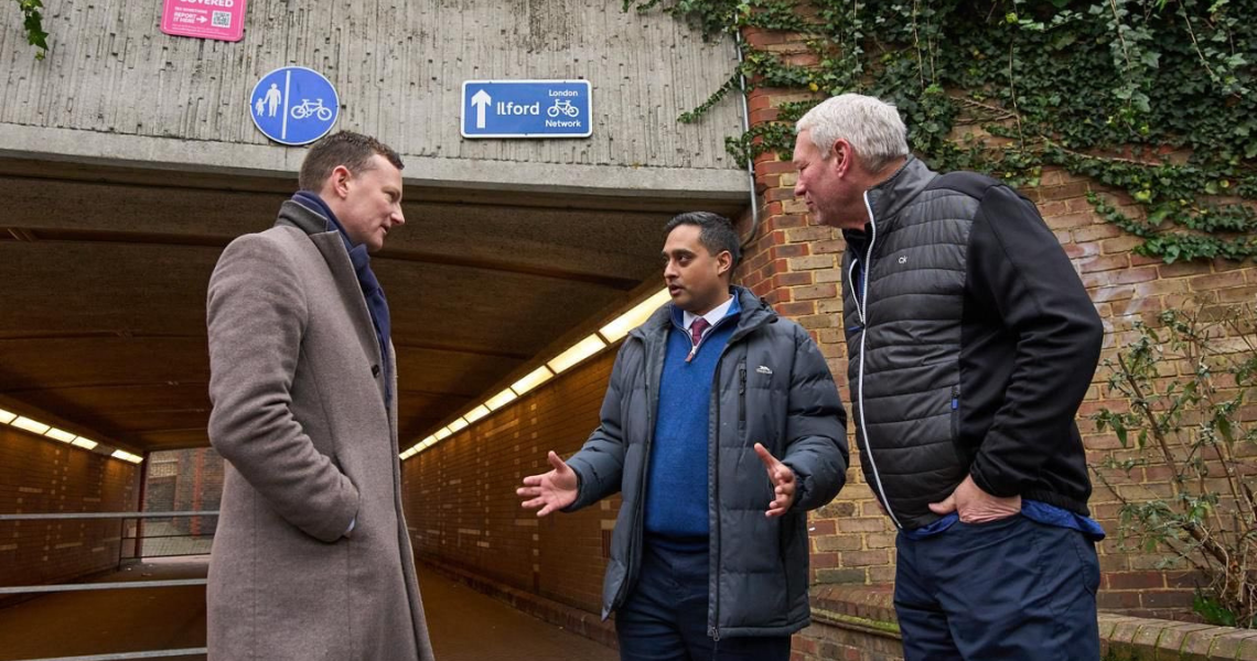 Three men engaged in conversation inside a tunnel located beneath a bridge, surrounded by concrete walls and dim lighting.